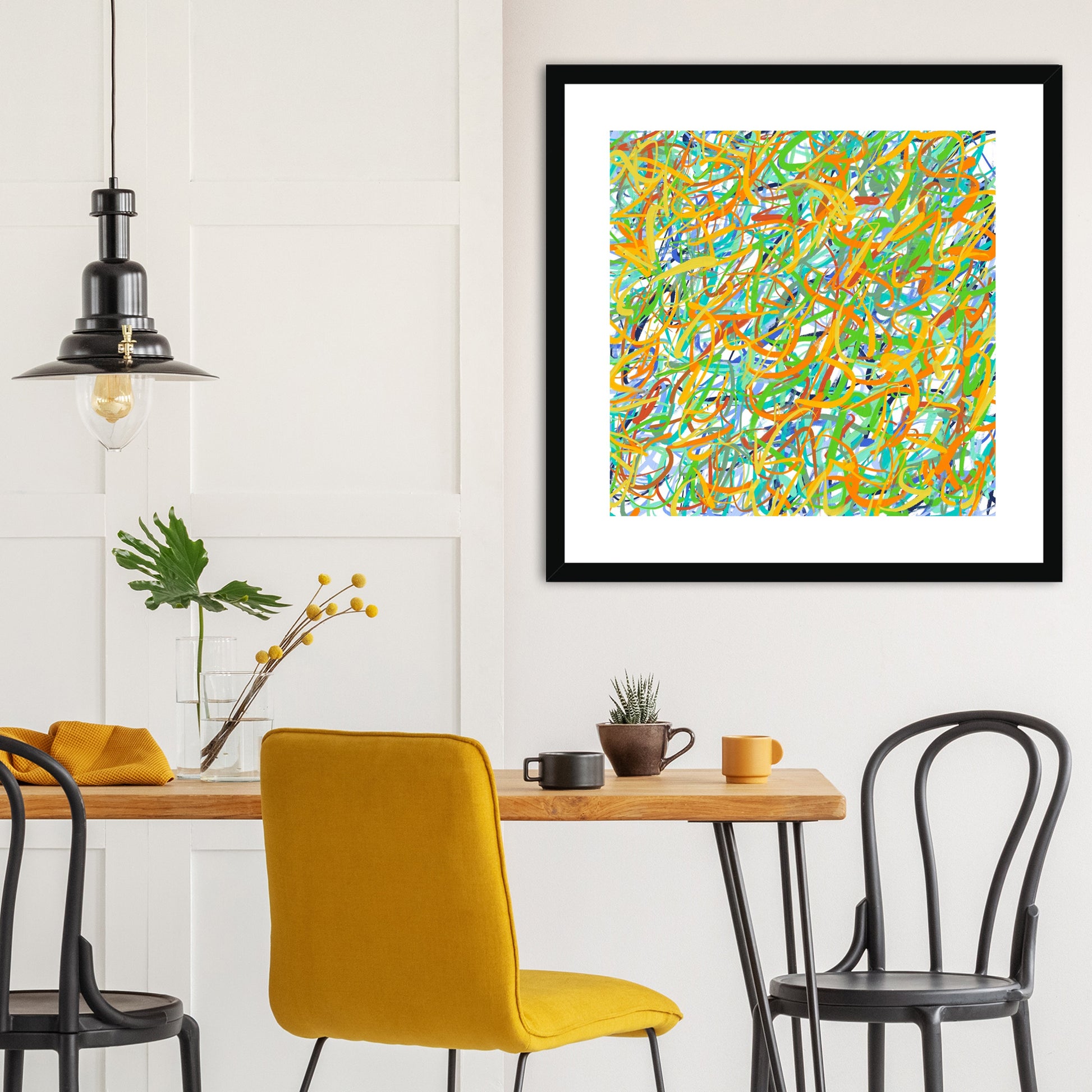 The black-framed artwork's tangle of yellows, greens and bluey-greens works perfectly in this dining area with wooden table-top, yellow and black dining chairs and accessories and with black industrial-style pendant lamp to one side.